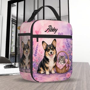 May include: A pink lunch bag featuring a watercolor print of a Corgi dog and a kitten in a basket amidst lavender flowers and hearts. The name Abby is printed in black script at the top. The bag has a black handle and a black mesh side pocket.