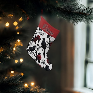 May include: A Christmas stocking with a red cuff and a design featuring black crows, red roses, and branches on a white background. The name "Olivia" is printed on the cuff. The stocking is hanging from a Christmas tree.