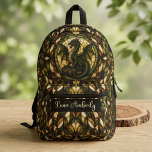 Puede incluir: Una mochila con un patrón de vidrieras en tonos verde, marrón y crema. El diseño incluye una ilustración central de un dragón verde. Una etiqueta rectangular negra en el bolsillo frontal muestra el nombre Luna Amberly en letras doradas.
