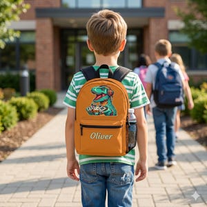 May include: Orange backpack featuring a green dinosaur wearing headphones and holding game controllers. The backpack has a black zipper pocket and a side mesh pocket for a water bottle. The name "Oliver" is printed on the front.