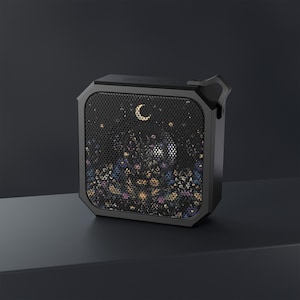 May include: A black portable speaker with a decorative front panel featuring a floral and celestial design. The speaker has a mesh grille with a crescent moon and star pattern. The speaker has a handle on the side.
