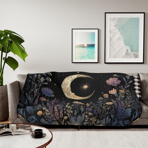 May include: A black throw blanket featuring a large, ornate golden crescent moon and a radiant star at the center, surrounded by a detailed botanical garden of blue, purple, and gold flowers, leaves, and stems against a dark night sky background.
