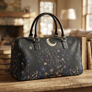 Puede incluir: Bolso de viaje negro con un diseño celestial que presenta una luna creciente, estrellas y elementos florales. El bolso tiene asas negras y cierre de cremallera. El bolso está sobre una superficie de madera.