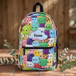 May include: A backpack featuring a colorful pattern of various kawaii-style cubes with smiling faces in shades of green, blue, purple, yellow, and pink. A light blue cloud-shaped patch in the center displays the name Emma in black text.