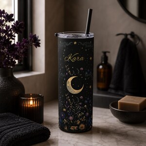 May include: A tall, cylindrical black tumbler featuring a gold crescent moon, scattered stars, and a colorful floral pattern at the base. The name Kara is printed in gold script near the top. The tumbler includes a clear lid and a black straw.