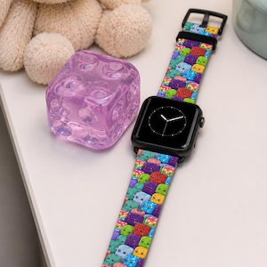 May include: A black smartwatch featuring a silicone band decorated with a repeating pattern of colorful, small, square-shaped cartoon characters with simple facial expressions in shades of blue, green, red, yellow, and purple.