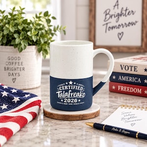 May include: A two-toned ceramic mug with a speckled white upper half and a solid navy blue lower half. The navy section features white text that reads CERTIFIED Talafreako 2026 A MORE SANE SENATE with star accents. The mug has a curved handle.