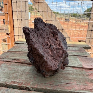 May include: A dark reddish-brown volcanic rock with a porous, vesicular texture. The stone features an irregular, jagged shape with numerous small holes and crevices across its surface, showcasing the natural cooling patterns of igneous rock.