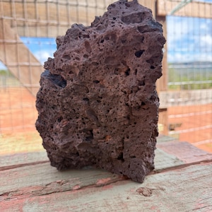 May include: A dark reddish-brown volcanic rock with a porous, vesicular texture. The stone features an irregular, jagged shape with numerous small holes and cavities across its surface, displaying a rough, natural appearance throughout the piece.
