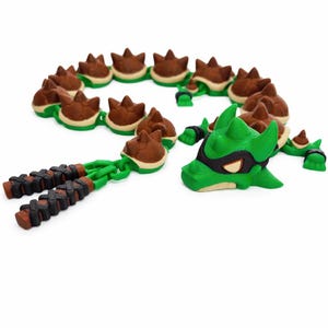 May include: A green and brown dragon-themed necklace and nunchucks set. The necklace features brown and tan spiked beads with green accents. The nunchucks have brown and black braided handles. A green dragon head with a black mask is also present.
