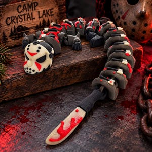 May include: A flexible, articulated toy figure shaped like a long, segmented creature with a hockey mask head, featuring a black and white color scheme with red accents. The tail end of the figure terminates in a pointed blade with red paint.