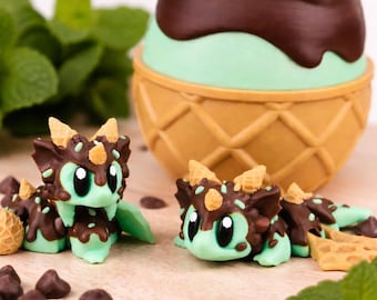 May include: A whimsical image features two mint green and chocolate brown dragon figurines with waffle cone accents. A large ice cream cone with a chocolate top sits in the background, along with chocolate chips and mint leaves.