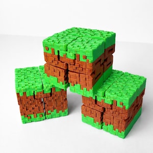 May include: Three green and brown cube-shaped toys, resembling blocks of grass and earth, are stacked on a white surface. The blocks are designed with a pixelated texture, reminiscent of a video game aesthetic.