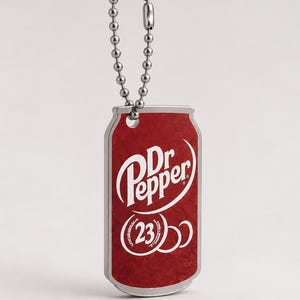 May include: A rectangular metal keychain shaped like a soda can with a silver ball chain. The front features a red background with the white Dr Pepper logo and the number 23 inside a circular graphic. The edges of the keychain are silver-toned metal.