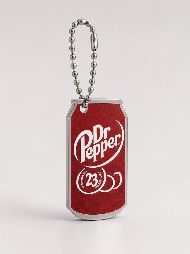 May include: A rectangular metal keychain shaped like a soda can with a silver ball chain. The front features a red background with the white Dr Pepper logo and the number 23 inside a circular graphic. The edges of the keychain are silver-toned metal.