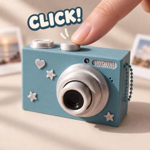 May include: A teal toy camera featuring a silver lens, a shutter button being pressed, and the text CLICK! above it. The camera body is decorated with small, glittery silver heart and star shapes, and it includes a matching teal ball chain.