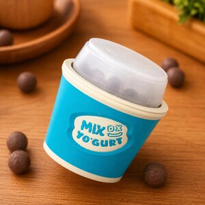 May include: A blue and white yogurt cup with the text "MIX YO'GURT" on the front. The cup is filled with small, round, dark brown candies. The cup is on a wooden surface, with more candies scattered around.