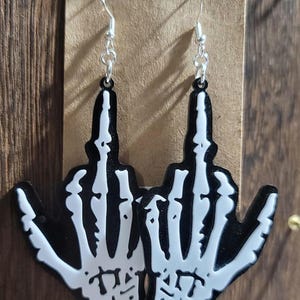 May include: A pair of black and white skeleton hand earrings. The earrings feature a detailed design of a hand with the middle finger extended, with a silver hook for the ear. The earrings are displayed on a brown background.
