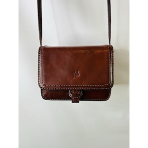 May include: A brown leather crossbody bag with a flap closure and a long, thin strap. The bag features a stitched design around the edges and a small emblem on the front. The bag is suspended against a white background.