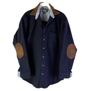 May include: A navy blue button-down shirt with brown suede elbow patches and a brown suede collar. The shirt has a chest pocket and is hanging on a white hanger. The cuffs are rolled up to reveal a light blue fabric.