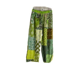 May include: Green patchwork pants with a drawstring waist. The pants feature a variety of green fabric patterns, including floral, paisley, and geometric designs. The pants are hanging on a white hanger.
