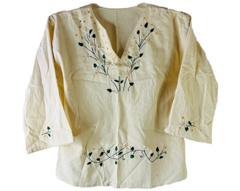 Puede incluir: Una blusa de manga larga de color crema con hojas verdes y flores blancas bordadas. El bordado está alrededor del cuello y en la parte inferior de la blusa.