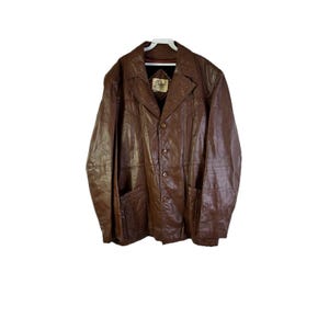 Peut inclure: Veste en cuir marron avec une fermeture boutonnée, des revers crantés et deux poches avant. La veste a un design classique et une esthétique vintage. Le cuir semble usé, ce qui ajoute à son caractère. La veste est suspendue à un cintre blanc.