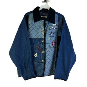 May include: A patchwork denim jacket with a black velvet collar. The jacket features various shades of blue denim, with floral and leaf embroidery in red, white, and blue. It has a button-up front and long sleeves.