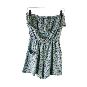 May include: A strapless floral print romper with a ruffled neckline and two pockets. The romper is a light blue color with a small floral pattern in shades of pink, purple, and yellow.