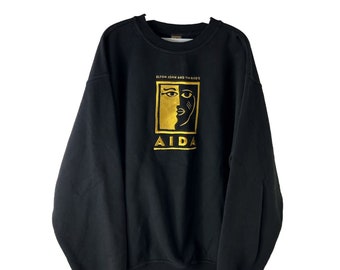 Puede incluir: Sudadera negra con un diseño bordado en dorado que presenta una cara estilizada y el texto "Elton John and Tim Rice's Aida".