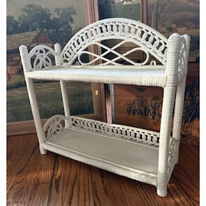 Può includere: Una mensola in vimini bianco a due livelli. Il ripiano superiore ha un design ad arco con tessitura decorativa. Il ripiano inferiore ha un bordo smerlato. La mensola è realizzata in vimini intrecciato e verniciata di bianco. È un oggetto decorativo per riporre oggetti.