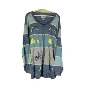 May include: A button-down cardigan sweater in shades of blue, lavender, and yellow. The sweater features embroidered flowers, butterflies, and dragonflies. The long-sleeved sweater has a V-neck and is hanging on a white hanger.