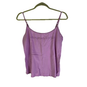 Puede incluir: Una camiseta sin mangas de color lila claro con tirantes finos, colgada de una percha verde. El texto "VICTORIA'S SECRET" está bordado en el pecho con hilo morado más oscuro. La parte superior tiene un escote redondo y un ajuste holgado.