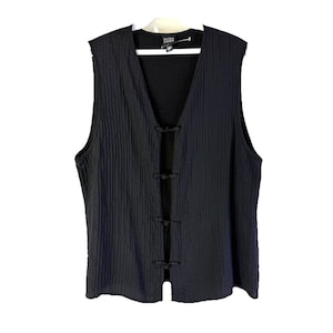 May include: A black sleeveless vest with a textured, crinkled fabric finish. The garment features a V-neckline and a front closure secured by three black fabric toggle knots. A white label with the text Eileen Fisher is visible at the inner neckline.
