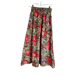 May include: A long, floral-print skirt with a patchwork design. The skirt features a red, green, and black color scheme with a variety of flowers. The skirt is hanging on a hanger.