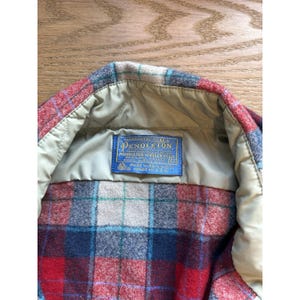 May include: Close-up of a Pendleton wool shirt. The shirt features a red, blue, and cream plaid pattern. The interior has a tan lining and a blue label with white text that reads "Pendleton Woolen Mills".