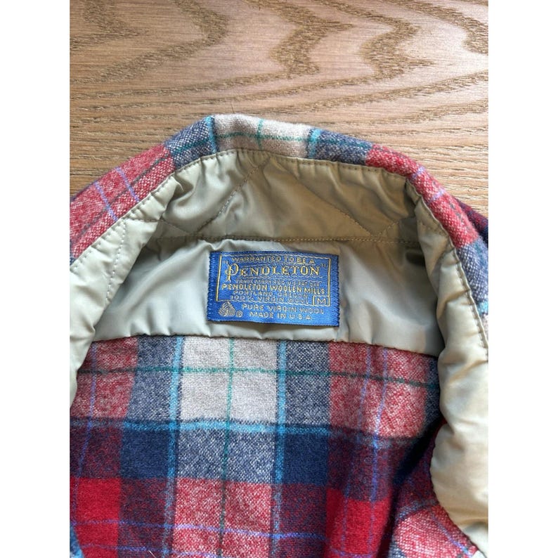 May include: Close-up of a Pendleton wool shirt. The shirt features a red, blue, and cream plaid pattern. The interior has a tan lining and a blue label with white text that reads "Pendleton Woolen Mills".