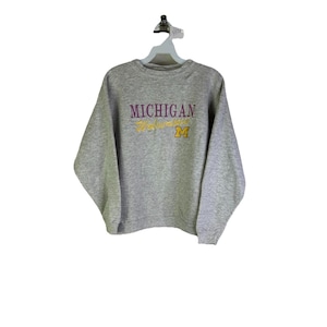 Op de afbeelding: Een grijs gemêleerde sweatshirt met het woord "MICHIGAN" in paarse blokletters en "Wolverines" in gouden letters. Een gouden "M"-logo staat eronder. De sweatshirt hangt aan een witte hanger.