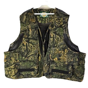 May include: Camouflage hunting vest with multiple pockets and a black mesh back and sides. The vest features a full-length zipper and a small logo patch near the collar. The pattern is a mix of greens, browns, and tans.