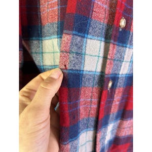May include: Close-up of a red, blue, and white plaid flannel shirt. The shirt has a button-down front and a classic plaid pattern. The fabric appears to be soft and warm, suitable for cooler weather.