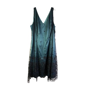 May include: A teal-colored formal dress with a V-neck and sleeveless design. The dress features a sheer overlay with dark floral embellishments, and a beaded hemline. The dress is suitable for special occasions.
