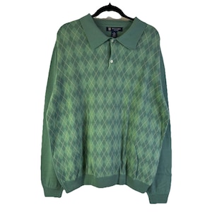 Può includere: Maglione verde a maniche lunghe con motivo a rombi. Il maglione ha un colletto e una chiusura con bottoni. Il marchio Oxford Golf è visibile sull'etichetta. Il maglione è appeso a una gruccia nera.