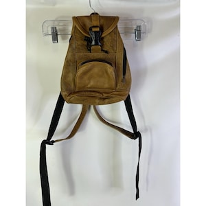 May include: A brown leather backpack with a flap closure and a front pocket. The backpack has black adjustable straps and a black buckle. The bag is hanging on a clear plastic hanger.