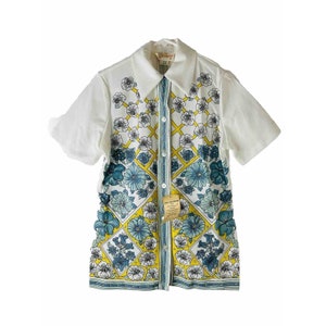 May include: A white short-sleeved button-down shirt with a floral pattern in shades of blue, yellow, and white. The shirt has a collar and a button closure. The pattern is a geometric design with squares and diamonds.
