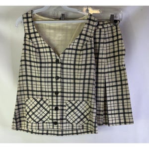 May include: A vintage two-piece outfit comprising a vest and a skirt. The vest has a V-neck, button closure, and two front pockets. Both items are cream-coloured with a black plaid pattern. The skirt features a pleated design.