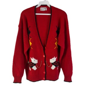 May include: A red cardigan sweater with a V-neck, featuring a button-up front and black trim. The sweater has white dog designs with black and red plaid accents, and yellow and black diamond patterns. The sweater is hanging on a white hanger.