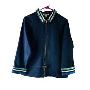 Puede incluir: Una chaqueta azul marino con cremallera, con cuello y puños a rayas blancas y verdes. La chaqueta tiene una cremallera dorada y una hebilla dorada en la cintura.