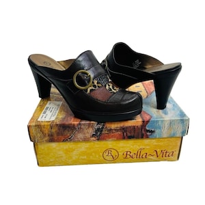 Op de afbeelding: Een paar zwarte leren sandalen met hak, een gesp en een veelkleurig patroon. De sandalen liggen op een doos met de tekst "By Bella Vita" erop.