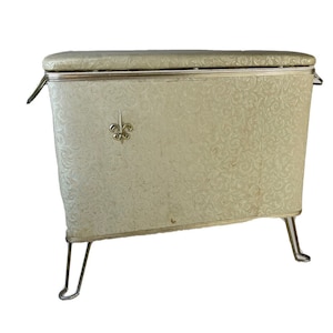 Könnte beinhalten: Ein vintage weißer Wäschekorb mit goldener Verzierung und einem Fleur-de-lis-Design. Der Korb hat einen gepolsterten Deckel und steht auf vier Metallbeinen.