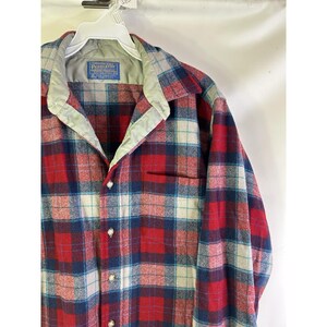 May include: A red, blue, and white plaid button-up shirt with a pocket. The shirt has a light green collar and a label that reads "Pendleton". The shirt is hanging on a white hanger.